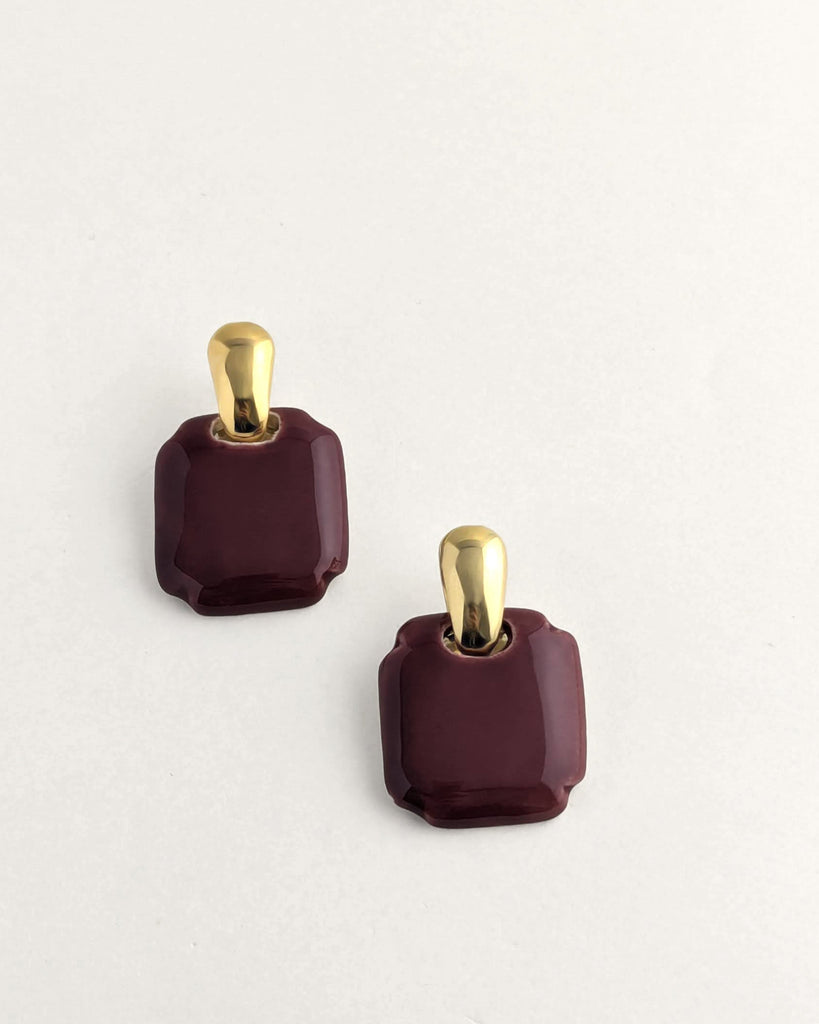 Paire de boucles d’oreilles forme carré arrondi coloris bordeaux, inspiration vintage année 80, au volume géométrique adouci, finition émaillée, montées sur un élément plaqué or.  Bijou contemporain fait main, style minimal et intemporel par Lydie Smith artisan bijoutier et céramiste basée à Paris.