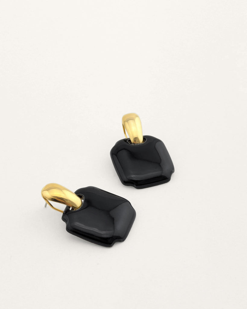 Paire de boucles d’oreilles forme carré arrondi coloris noir, inspiration vintage année 80, au volume géométrique adouci, finition émaillée, montées sur un élément plaqué or.  Bijou contemporain fait main, style minimal et intemporel par Lydie Smith artisan bijoutier et céramiste basée à Paris.