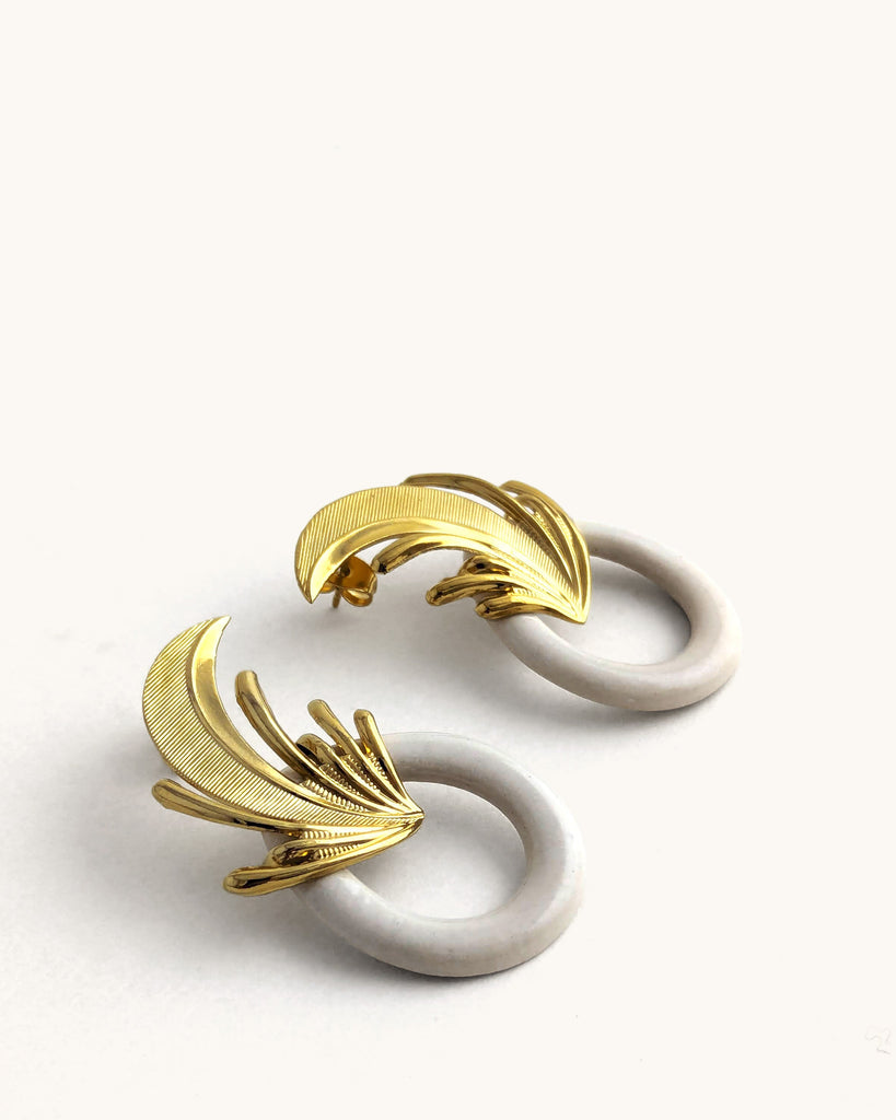 Boucles d’oreilles art déco blanc crème et dorées, ornées d’un motif feuille sculpté rappelant l’élégance des années Gatsby, avec un anneau noir laqué contrastant. Créé par LYDIE SMITH artisan céramiste et bijoutier dans son atelier à Asnières en France
