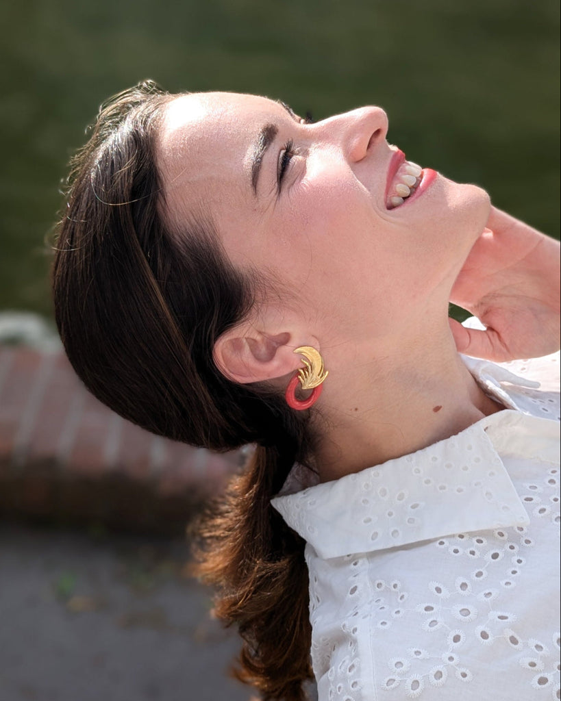 Femme heureuse portant les boucles d’oreilles art déco rouge et dorées, ornées d’un motif végétal sculpté rappelant l’élégance des années Gatsby, avec un anneau noir laqué contrastant. Créé par LYDIE SMITH artisan céramiste et bijoutier dans son atelier à Asnières en France